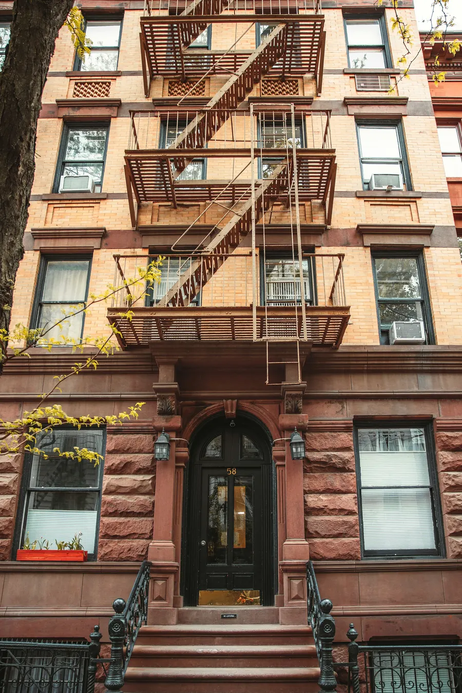 A New York brownstone with a black fire escape climbing its pale brick front above an arched doorway