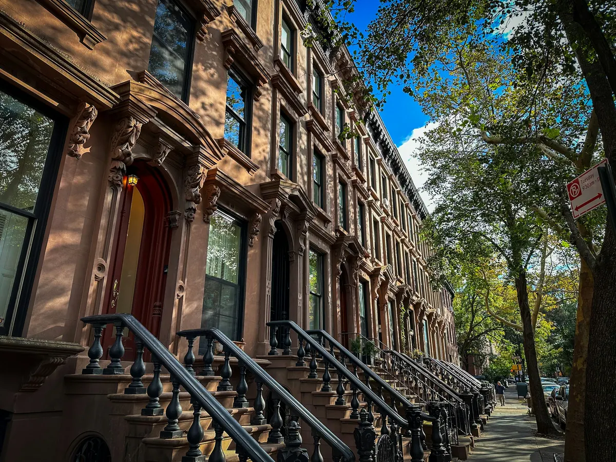 A tree-lined New York street of brownstone houses with black iron stair railings in dappled light