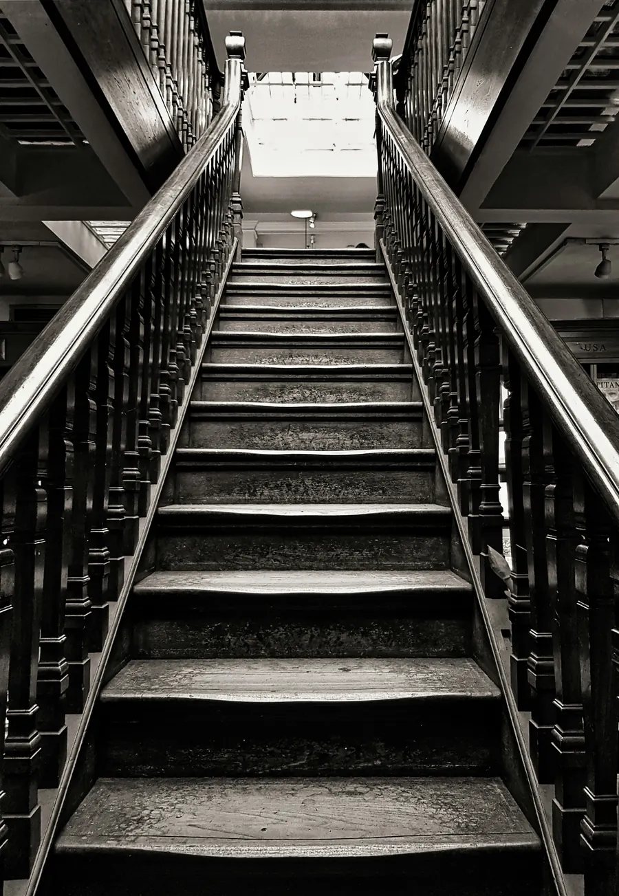 A steep wooden staircase with dark turned balusters and polished handrails rising toward a bright skylight, in black and white
